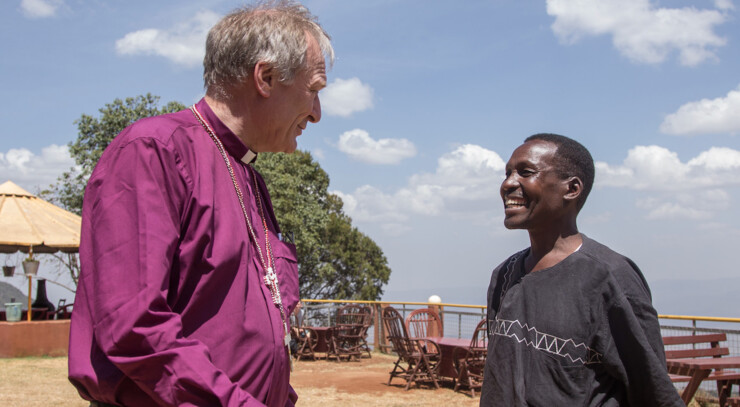 The image depicts Bishop Henrik Stukjær engaged in conversation. Henrik Stubkjær is wearing a purple robe, while the other individual is dressed in black attire with white patterns on the sleeves. The background scene features wooden chairs and tables under an umbrella, suggesting an outdoor seating area. The sky is clear with a few clouds, indicating a sunny day.