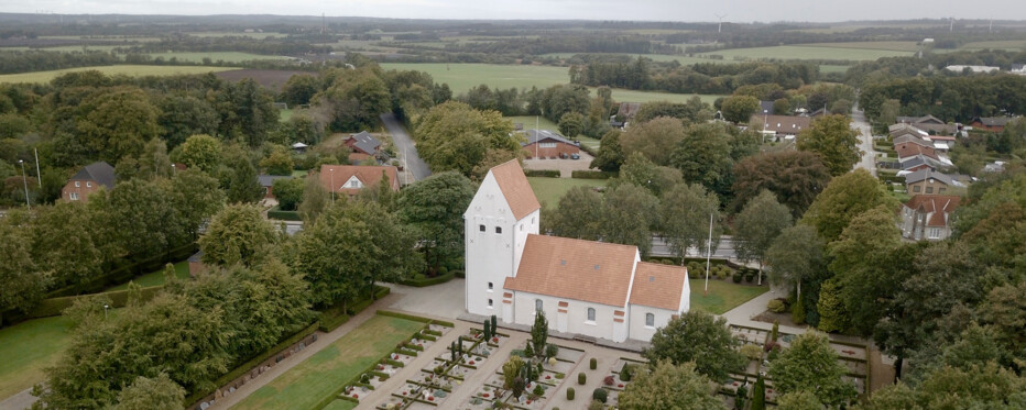 Billedet viser et luftfoto af Haunstrup kirke, der er omgivet af grønne træer og huse. Kirken har en markant hvid struktur med et stejlt, spidst tag. Foran kirken er der flere grave med gravsten, der er organiseret i rækker, hvilket indikerer, at det er en kirkegård. Området omkring kirken er grønt med træer spredt mellem boligbygningerne. En vej strækker sig fra forsiden af kirken til højre side af billedet og fører ind i et nabolag med flere synlige huse. Himlen er overskyet og kaster et blødt lys over hele scenen.