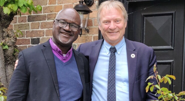 Musa Panti Filibus, along with Henrik Stubkjær, both dressed in suits, stands side by side in front of a brick wall and a black door. Above them hangs an outdoor lamp attached to the wall.